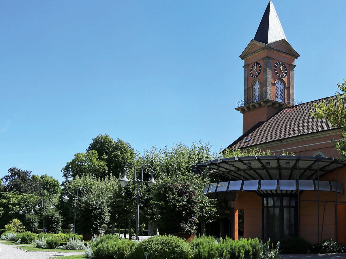 Historische Kirche mit markantem Uhrturm und modernem Anbau in einer grünen Parkanlage an einem sonnigen Tag.