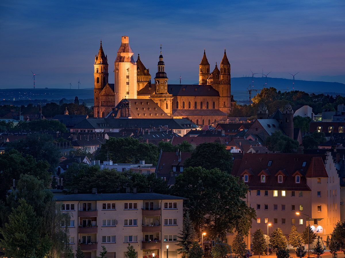 Dom zu Speyer bei blauer Stunde, beleuchtet über der Stadt mit Häusern, Bäumen und Windrädern im Hintergrund.