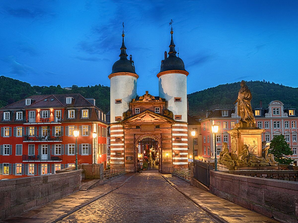 Alte Brücke Heidelberg bei Nacht: beleuchtetes Brückentor, historische Gebäude, Karl-Theodor-Statue, blauer Abendhimmel.