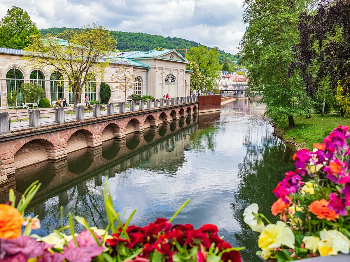 Historische Architektur am Fluss Oos in Baden-Baden mit Personen auf Promenade, grünen Bäumen und bunten Blumen im Frühling.