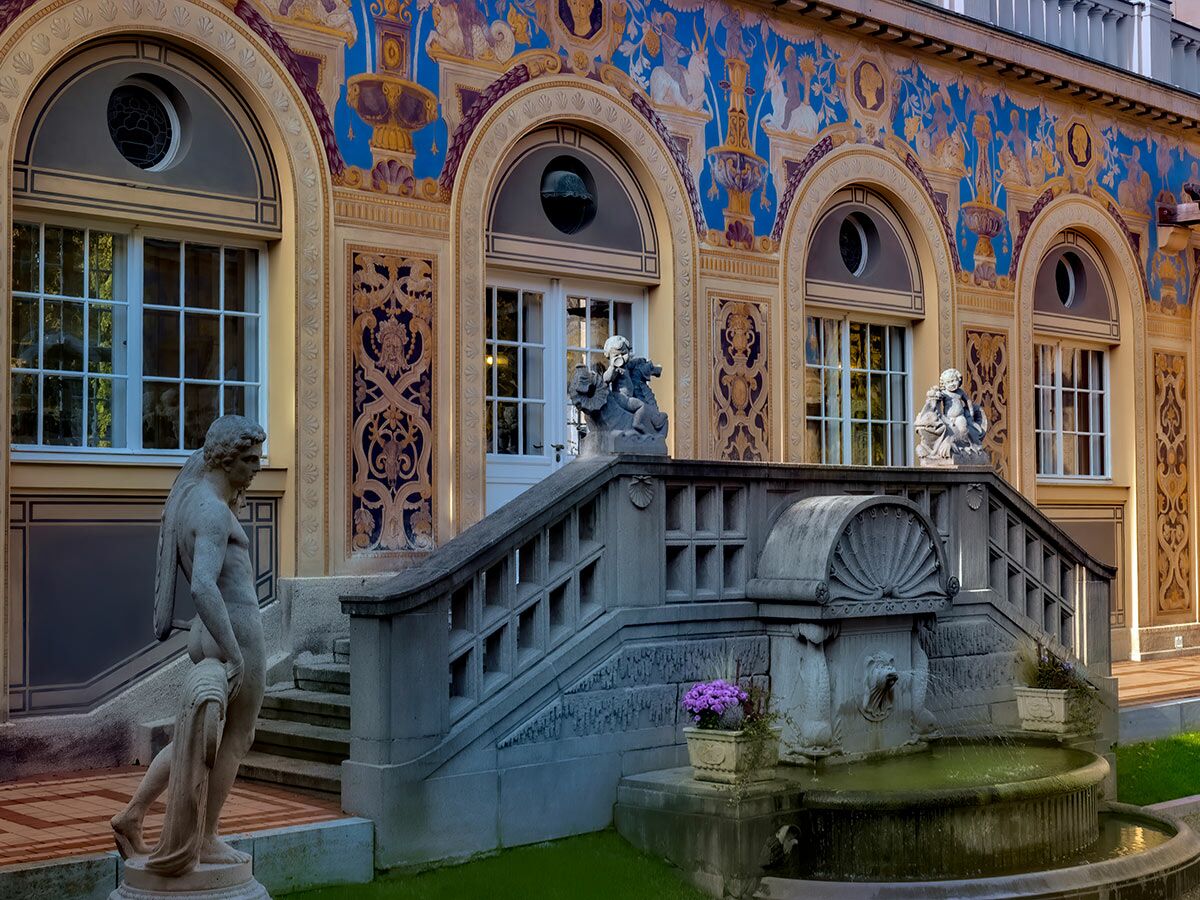 Ornate historische Gebäudefassade mit blau-goldenen Wandmalereien, Steintreppe, Skulpturen und einem Brunnen im Garten.