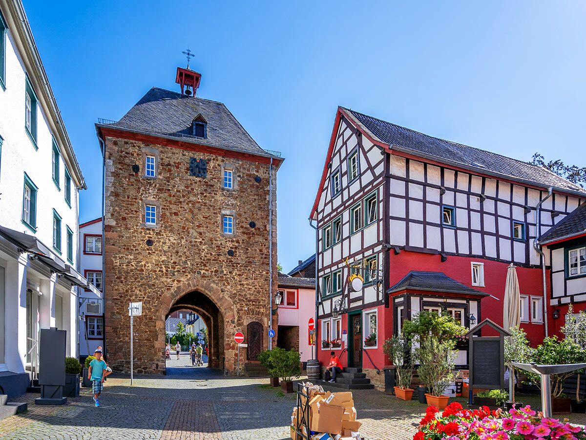 Sonnenbeschienener Marktplatz mit mittelalterlichem Torbogen, rot-weißem Fachwerkhaus und Personen in historischer deutscher Stadt.