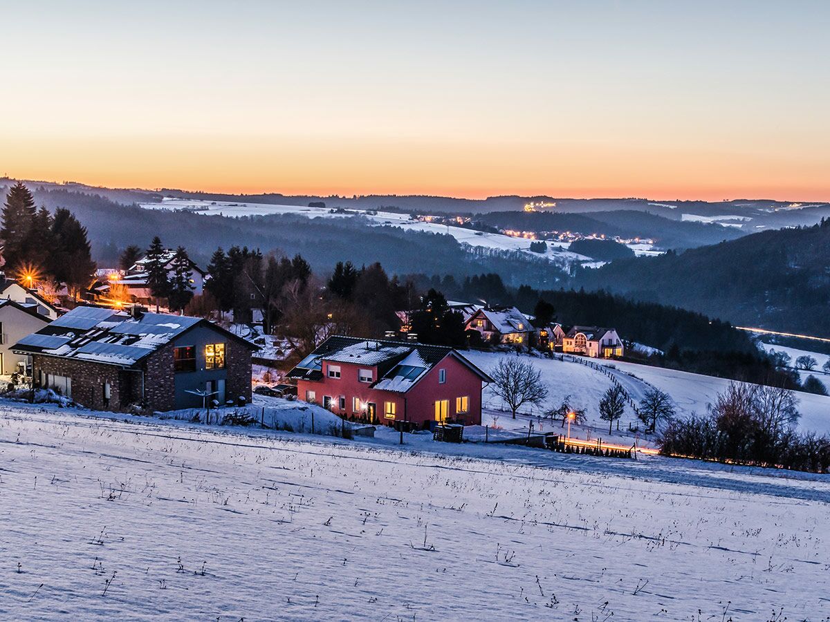 Idyllische Winterlandschaft bei Sonnenuntergang mit schneebedeckten Hügeln, beleuchteten Häusern und warmem Abendlicht.
