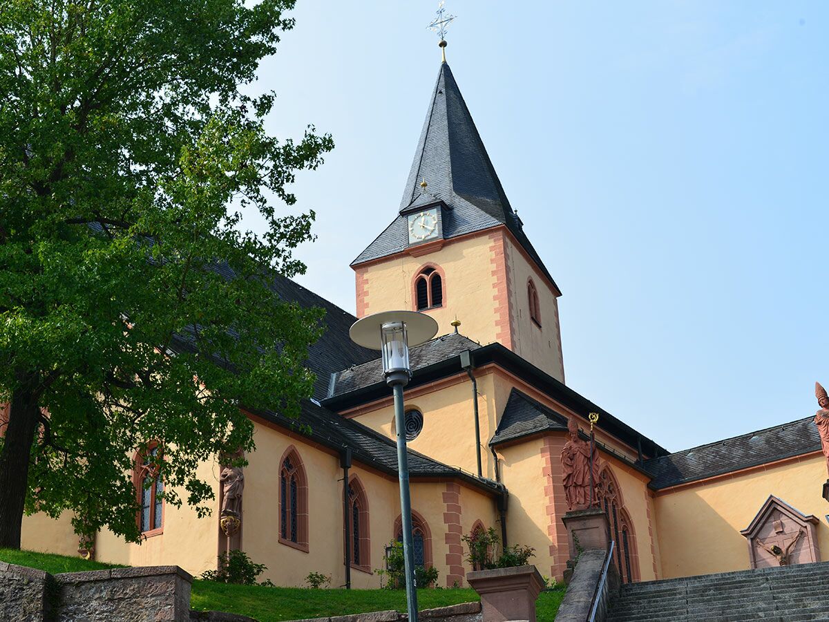 Alte Kirche mit spitzem Turm, Uhr, roten Sandsteinakzenten, Statuen, großem Baum und Treppe unter blauem Himmel.