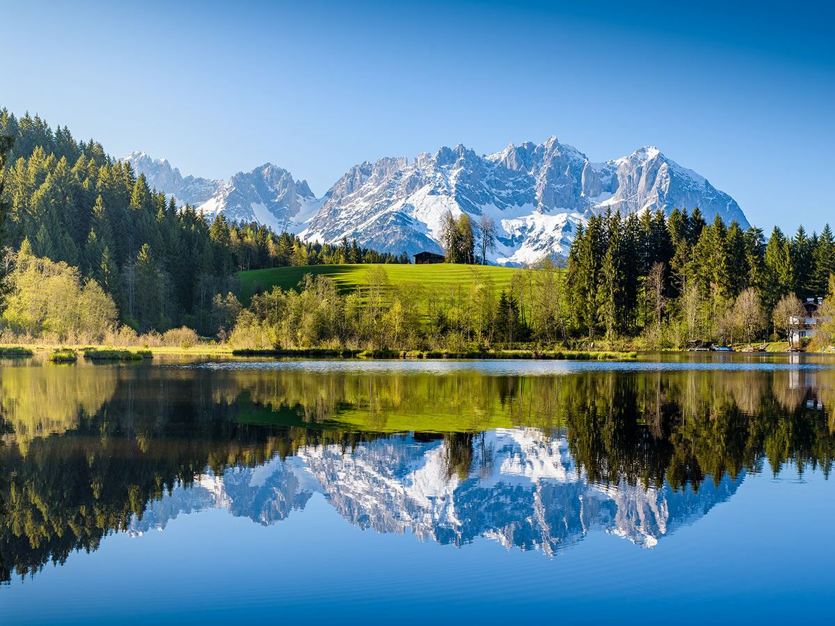 Idyllische Alpenlandschaft mit schneebedeckten Bergen, grünen Wäldern und einem klaren See, der die Landschaft spiegelt.