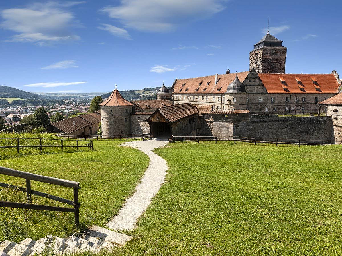 Panoramablick auf die Plassenburg in Kulmbach, Bayern. Eine historische Burg mit Türmen, roten Dächern und einer grünen Wiese im Vordergrund, dahinter die Stadt Kulmbach und Hügel unter blauem Himmel.