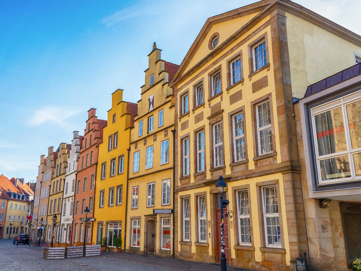 Reihe bunter, historischer Giebelhäuser in einer Altstadtstraße unter blauem Himmel. Zeigt detaillierte Fassaden und Fenster.