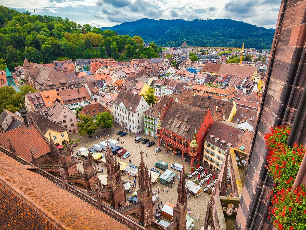 Luftaufnahme Münsterplatz Freiburg: belebter Markt mit Ständen, historischen Häusern, Minster-Details, grünen Bergen im Hintergrund.