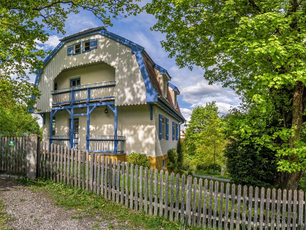 Charmantes blau-weißes Haus mit Balkon und markantem Dach, umgeben von grünen Bäumen und einem Holzzaun.