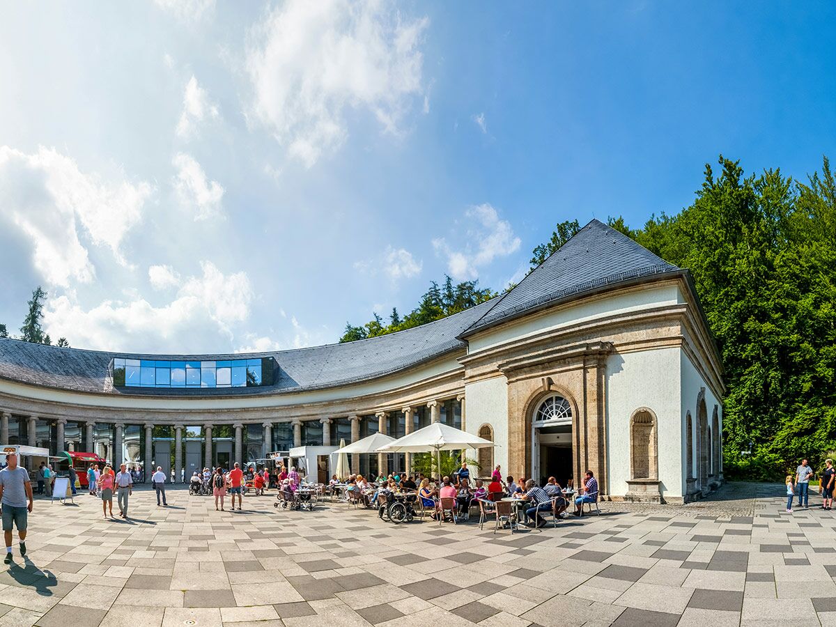 Panoramablick auf die Wandelhalle Bad Brückenau mit Menschen im Freien, Café-Terrasse und historischer Architektur.