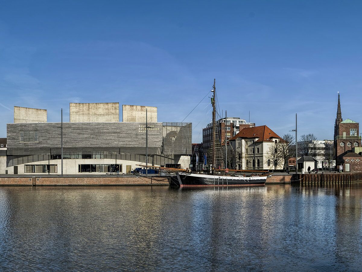 Das Deutsche Auswandererhaus in Bremerhaven am Hafen, mit Segelschiff und historischen Gebäuden unter blauem Himmel.