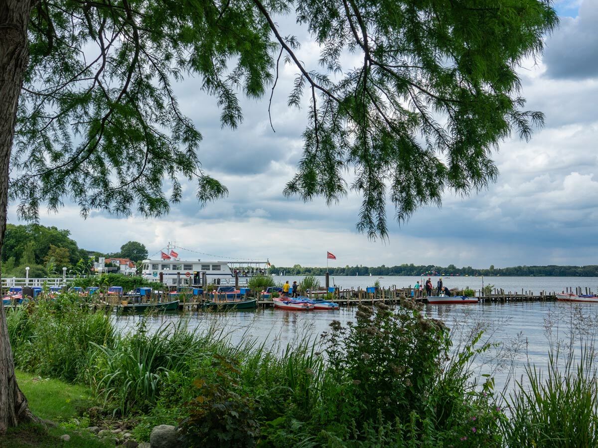 Seeufer mit Holzpier, Passagierschiff, Mietbooten und Menschen unter bewölktem Himmel, umgeben von grüner Natur.