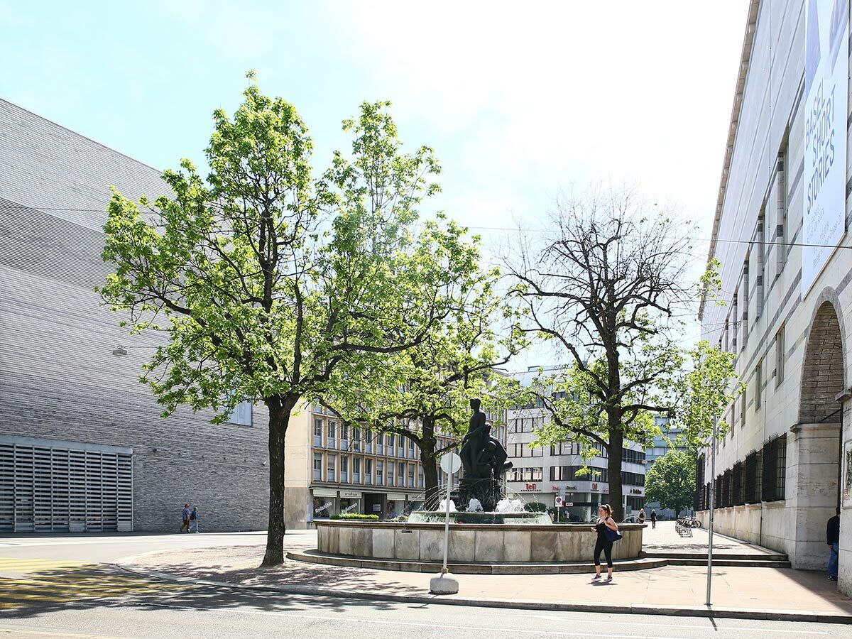 Sonniger Stadtplatz in Basel mit Skulpturenbrunnen, verschiedenen Bäumen und einer Mischung aus moderner und klassischer Architektur.