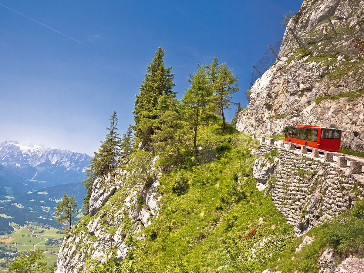 Roter Reisebus fährt auf einer schmalen Bergstraße mit steilen Felsen und Bäumen, Blick ins Tal und auf Schneegipfel.
