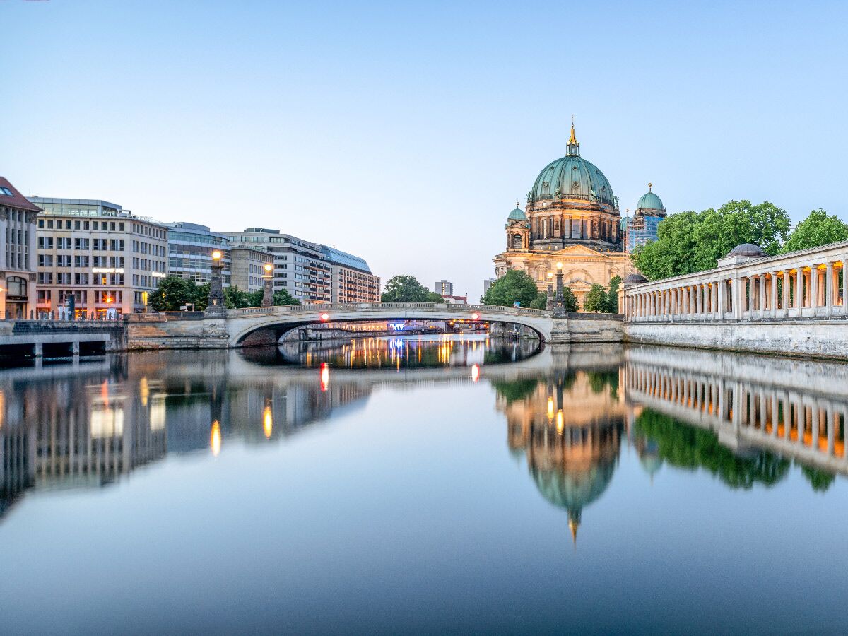 Der Berliner Dom beleuchtet bei Sonnenuntergang an der Spree, mit Brücke und Gebäuden, Spiegelung im ruhigen Wasser.