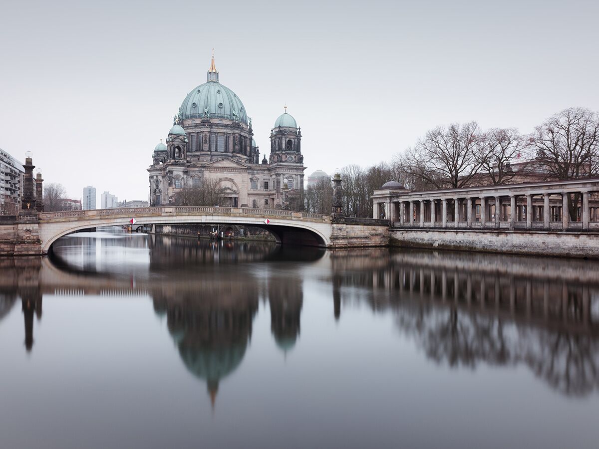 Panoramablick auf den Berliner Dom und eine Bogenbrücke über die Spree, gespiegelt im ruhigen Wasser an einem bewölkten Tag.
