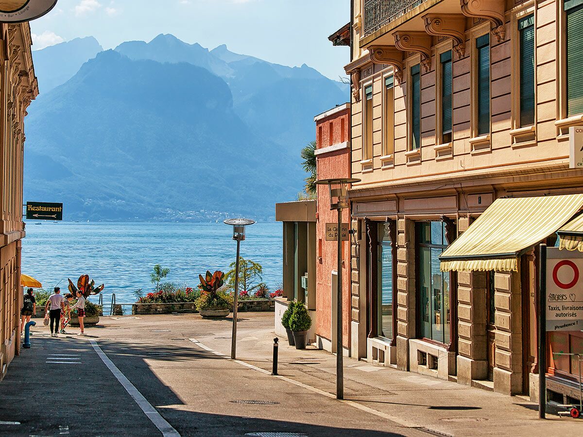 Sonnige Seepromenade in der Schweiz mit Fußgängern, historischen Gebäuden und klarem See- und Bergpanorama.