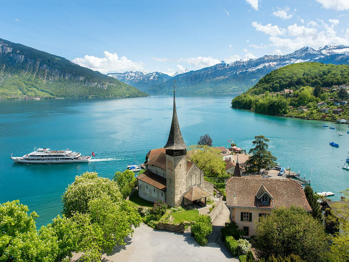 Blick auf die Iseltwald Kirche am Brienzersee mit einem Passagierschiff, grünen Bäumen und schneebedeckten Alpen unter blauem Himmel.