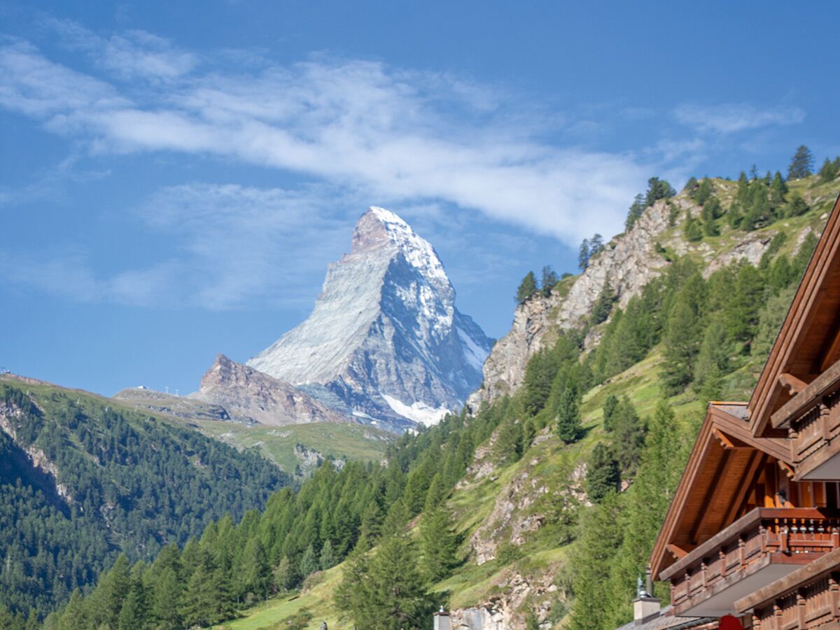 Majestätisches Matterhorn mit schneebedeckter Spitze, umgeben von grünen Berghängen und Wald, blauer Himmel über Zermatt.