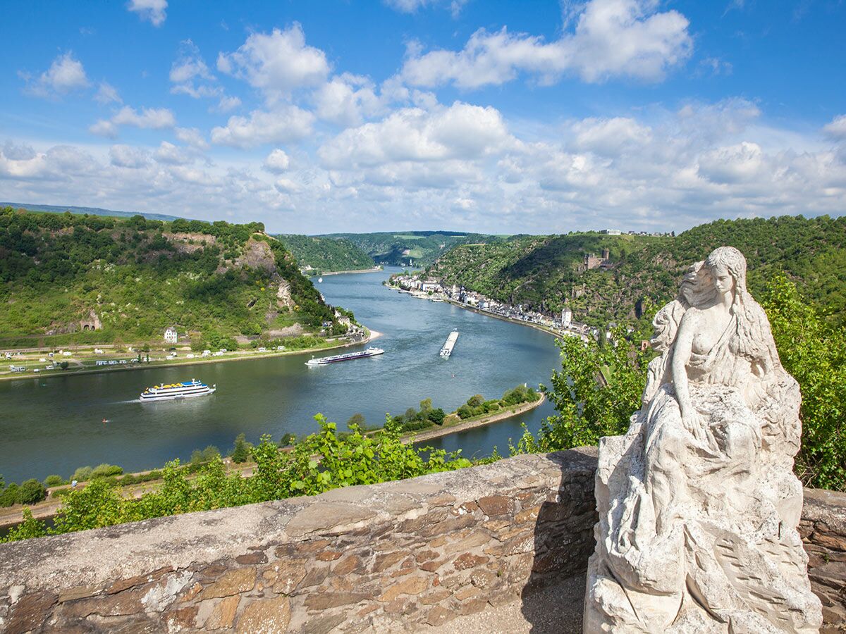 Panorama des Rheintals mit Loreley-Statue, Fluss, grünen Hügeln, Dorf und Schiffen unter blauem Himmel mit Wolken.