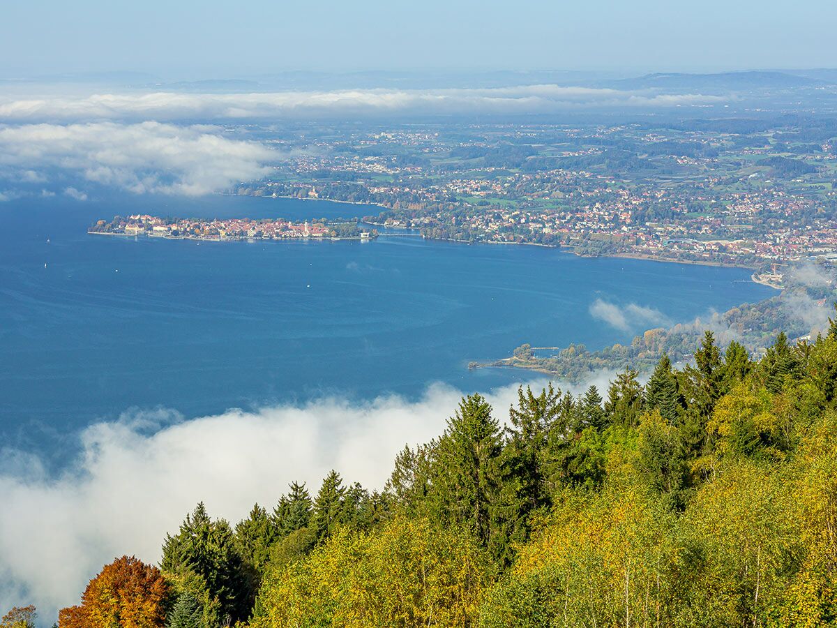 Panoramablick auf die Inselstadt Lindau im Bodensee, umgeben von Herbstbäumen und Nebel über dem Wasser.