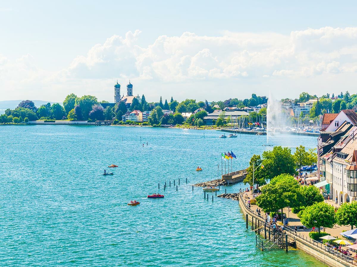 Blick auf eine lebhafte Seeuferpromenade und historische Stadt mit markanter Kirche an einem sonnigen Sommertag. Menschen genießen Wassersport.