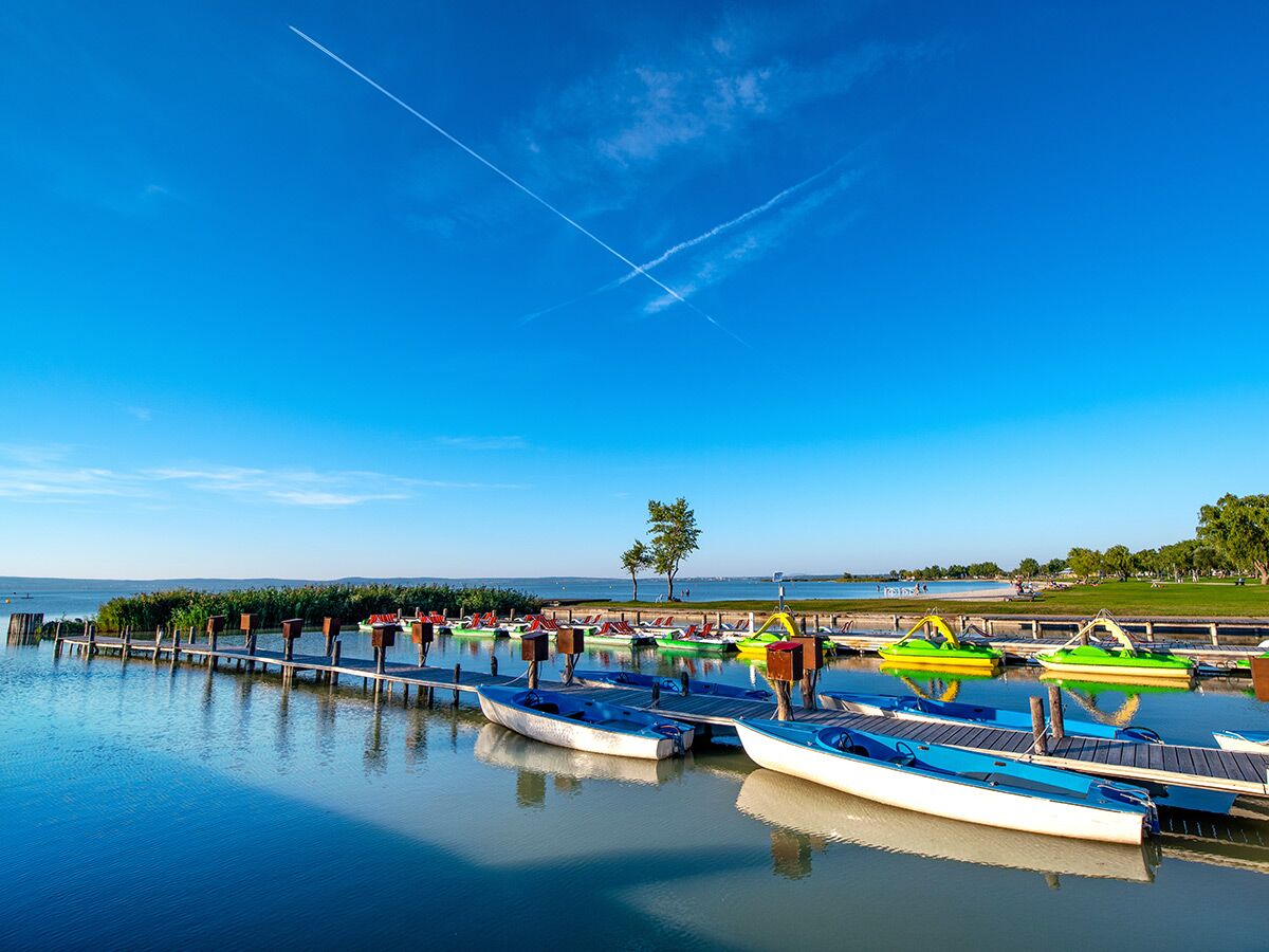 Sonniger Tag am See mit blauen und weißen Tretbooten sowie farbigen Paddelbooten an einem Holzsteg. Klarer blauer Himmel.