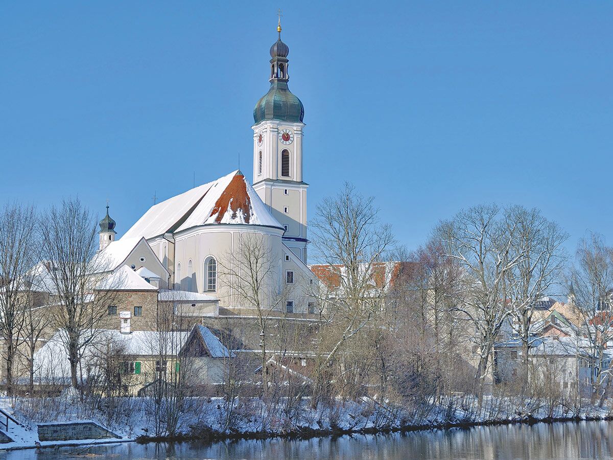 Barocke Kirche mit hohem Glockenturm und schneebedeckten Dächern an einem Fluss im Winter unter blauem Himmel.