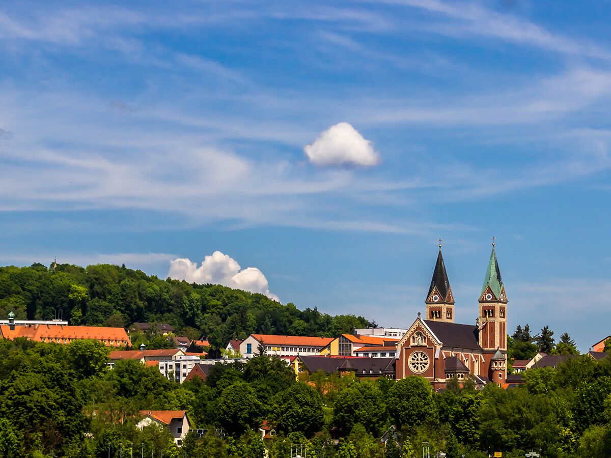 Imposante Kirche mit zwei markanten Türmen und roten Dächern inmitten einer grünen Hügellandschaft unter blauem Himmel mit Wolken.