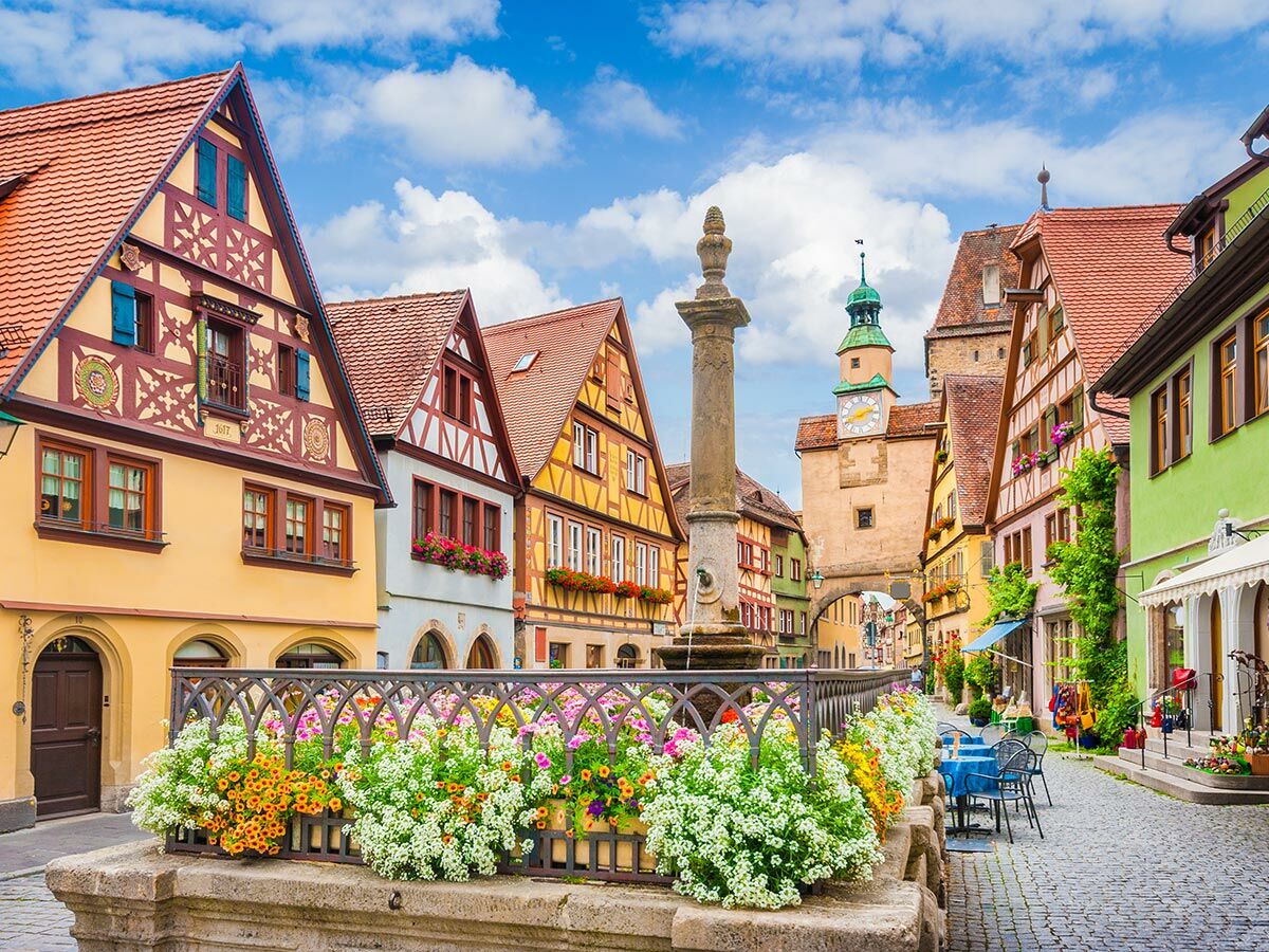 Malerische Altstadtgasse mit farbigen Fachwerkhäusern, blühenden Blumen am Brunnen und historischem Uhrturm unter blauem Himmel.