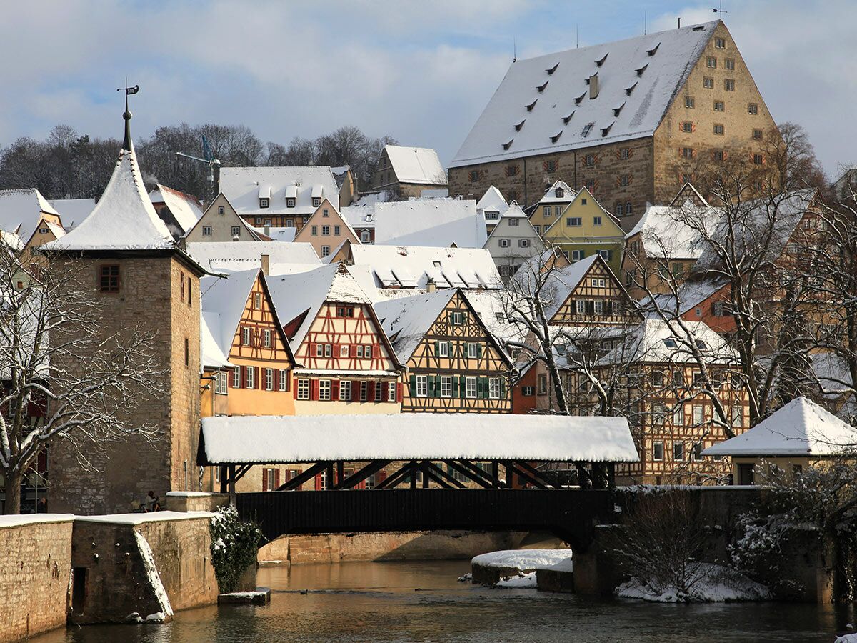Schneebedeckte Fachwerkhäuser, historischer Turm und überdachte Holzbrücke über dem Kocher in Schwäbisch Hall, Deutschland.