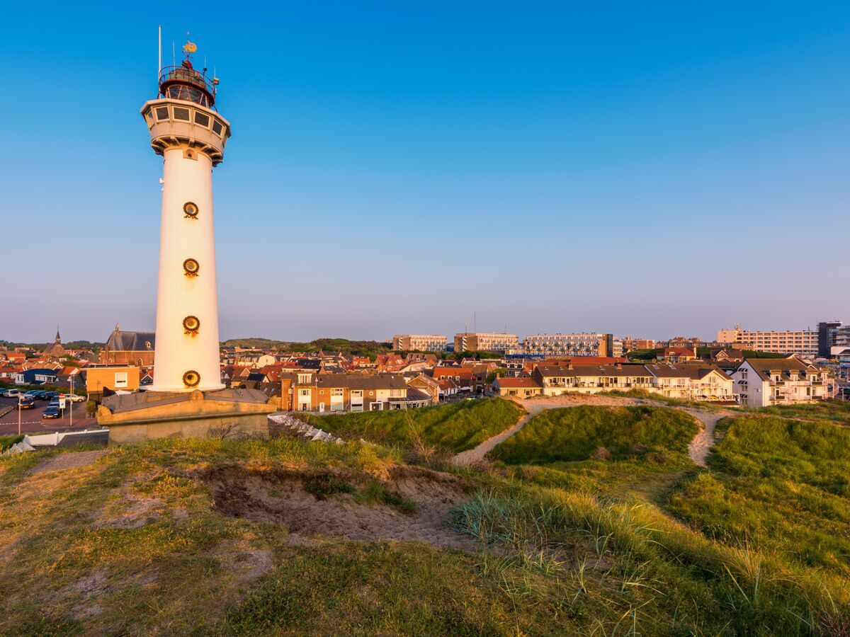 Hoher, weißer Leuchtturm auf einer Düne, überblickend eine malerische Küstenstadt unter blauem Himmel am Abend.