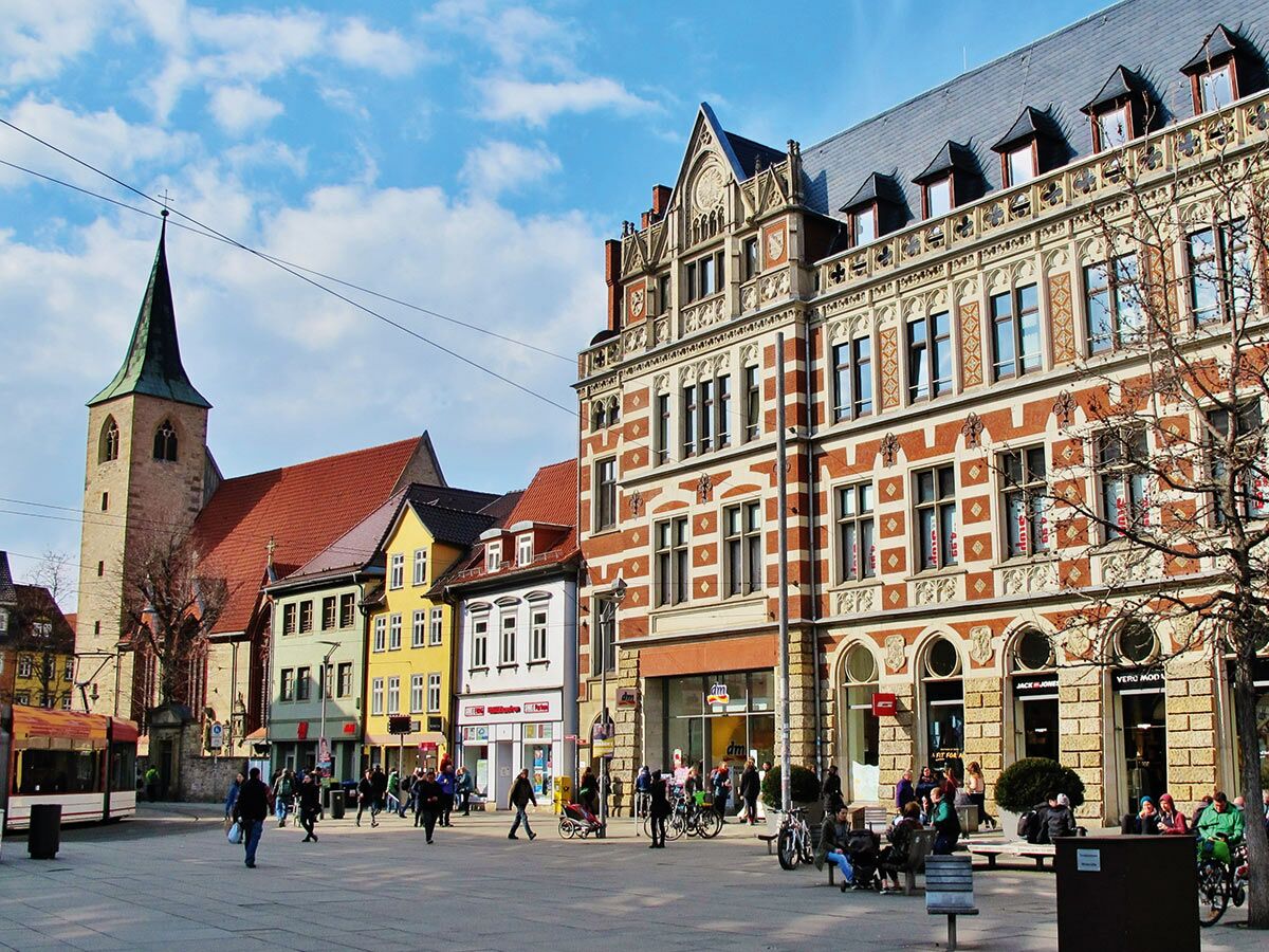 Ein belebter Stadtplatz in einer historischen deutschen Stadt, mit einer Kirche, alten Gebäuden, Geschäften, vielen Menschen und einer Straßenbahn.
