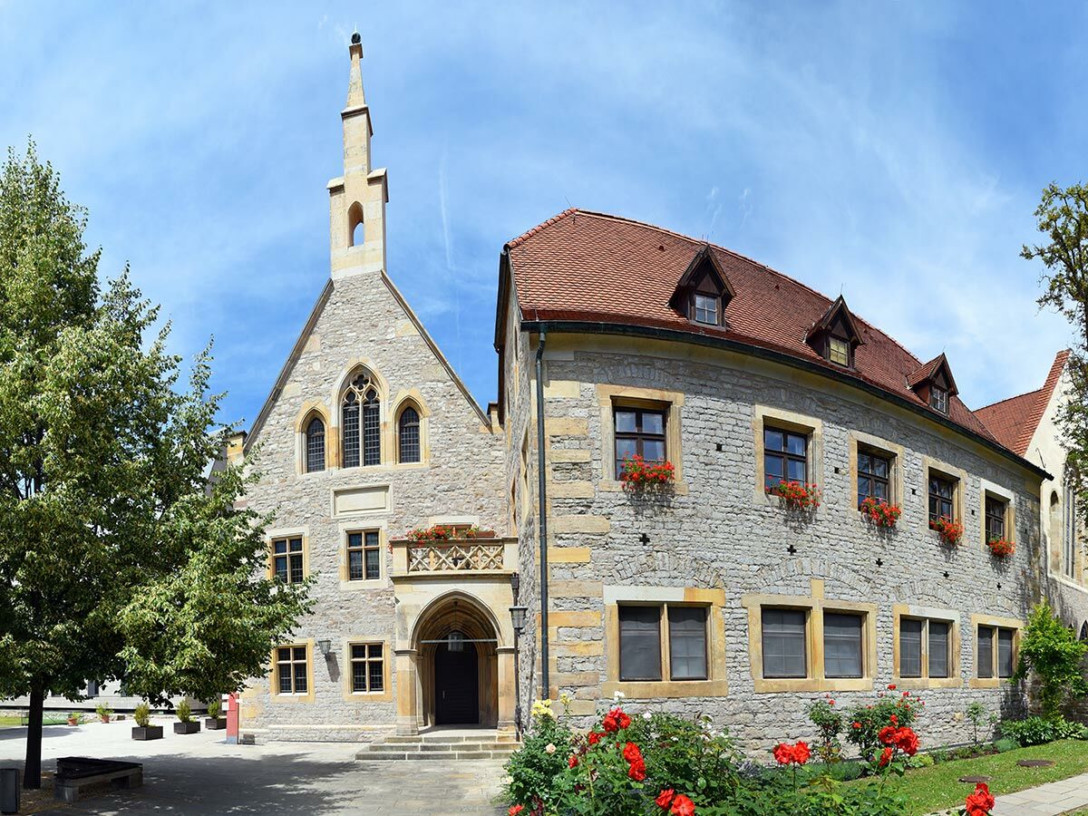 Historisches Steingebäude der Domus Universitatis Jena mit spitzem Turm, gotischen Fenstern und rotem Dach unter blauem Himmel, umgeben von Bäumen und Rosen.