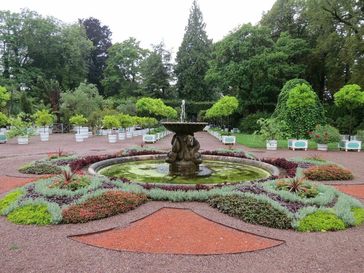 Üppige Gartenanlage mit zentralem, steinernem Brunnen, umgeben von kunstvoll angelegten Blumenbeeten und Kieswegen.