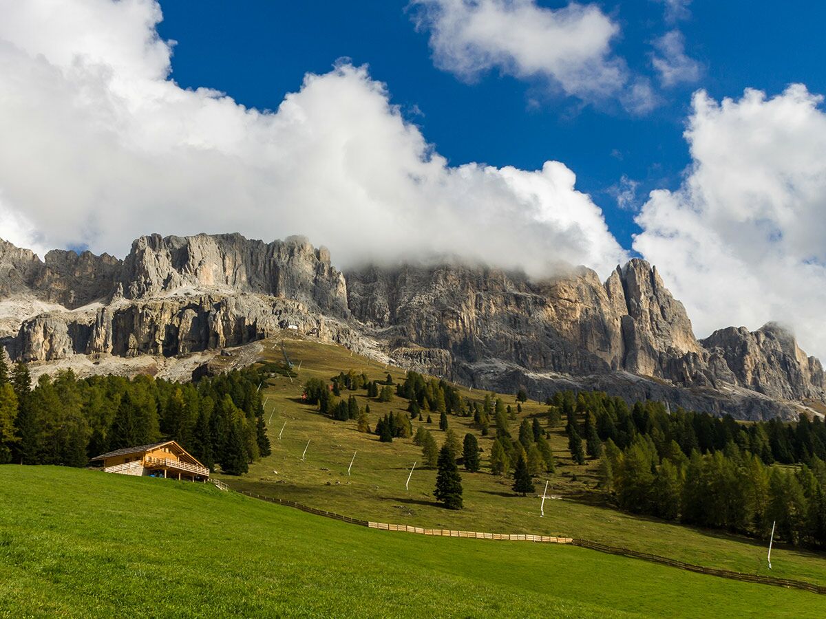 Majestätische Dolomiten mit einem traditionellen Holzchalet auf einer grünen Wiese unter blauem Himmel und weißen Wolken.