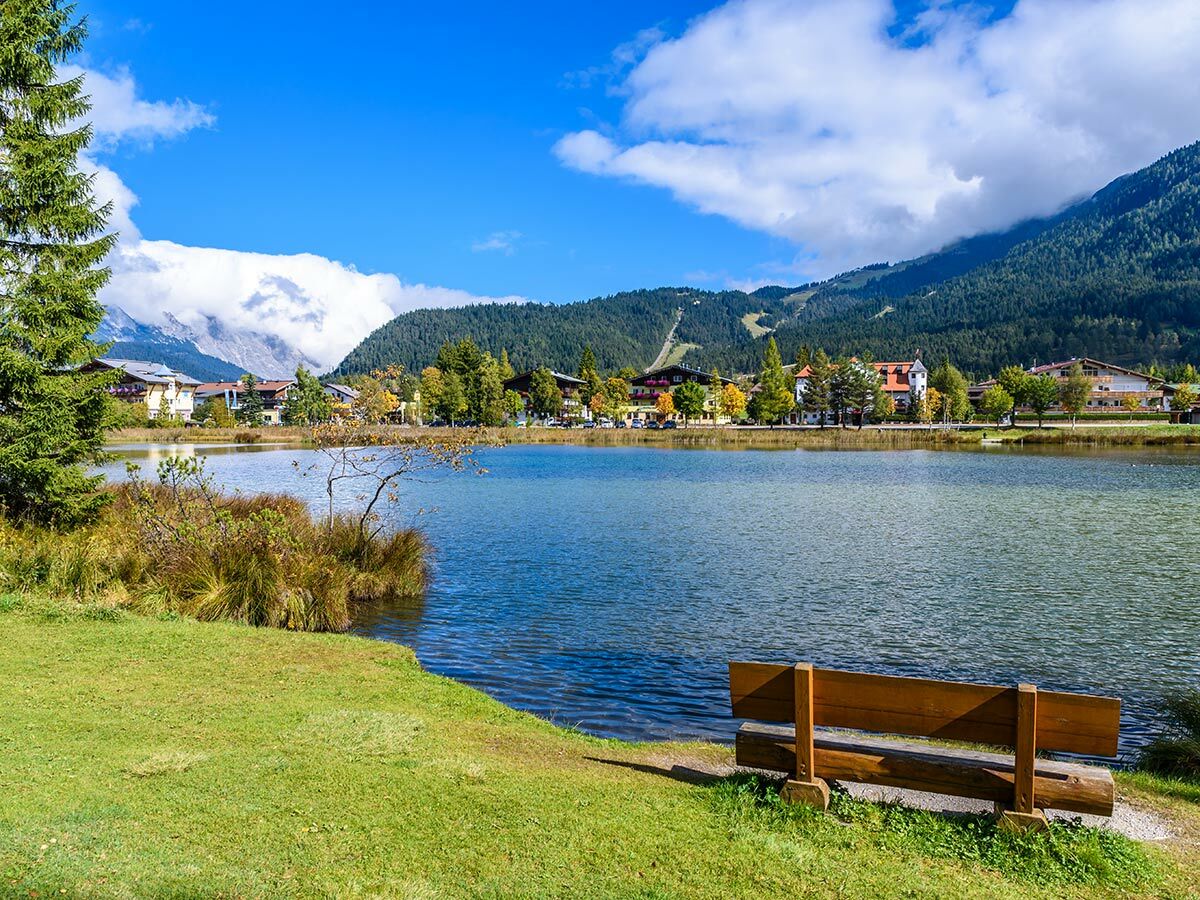 Panoramablick auf einen idyllischen Alpen See mit blauem Himmel, bewaldeten Bergen, einem Dorf am Ufer und einer Holzbank im Vordergrund im Herbst.