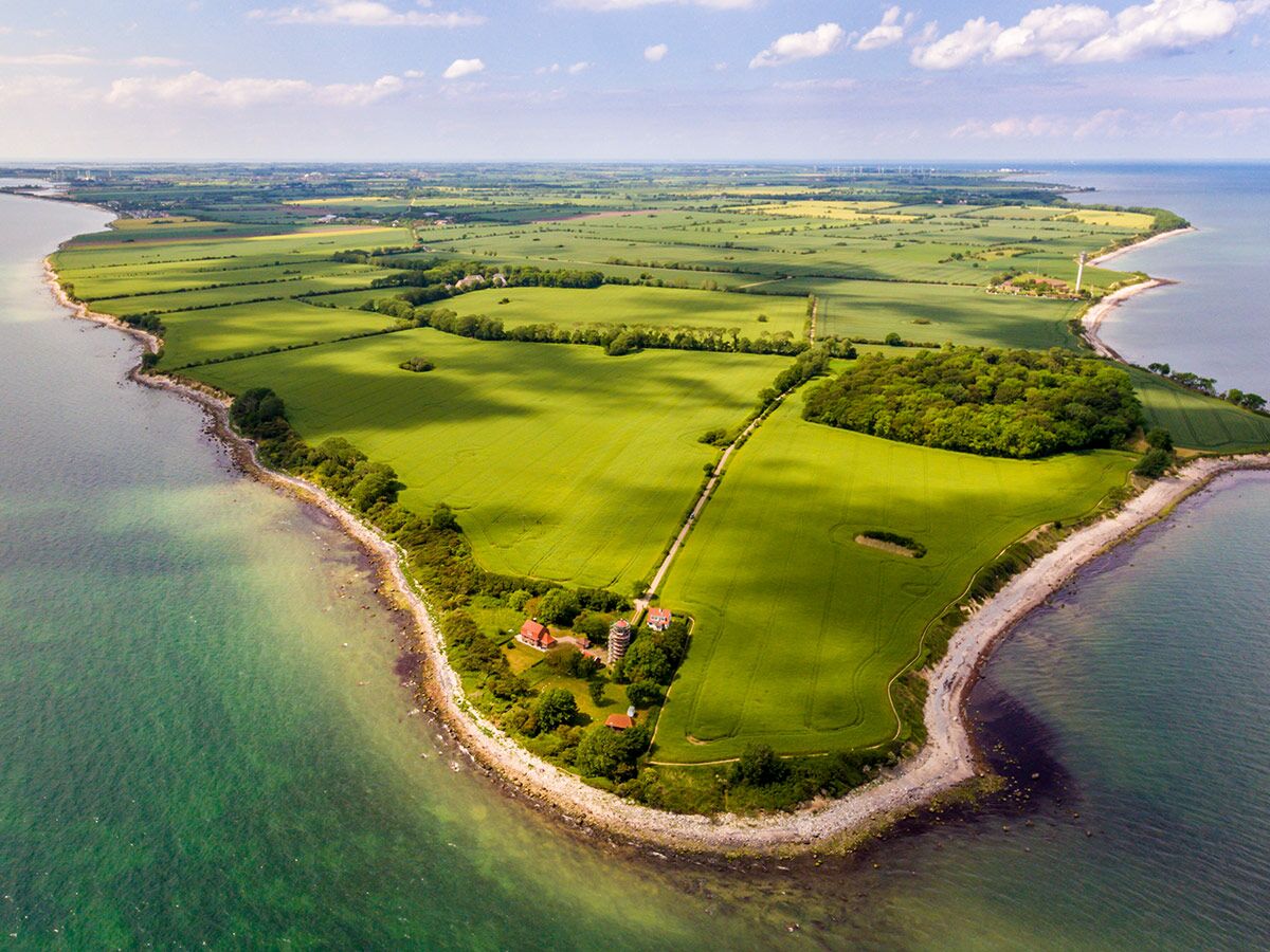 Luftaufnahme einer malerischen Küstenlandschaft mit grünen Feldern, blauem Meer, kleinen Wäldern und einem Leuchtturm bei Sonnenschein.