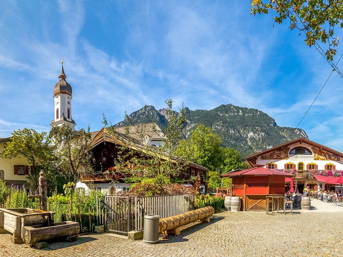 Malerisches bayerisches Dorf mit Kirchturm, traditionellen Häusern und belebtem Platz vor majestätischem Alpenpanorama bei Sonnenschein.