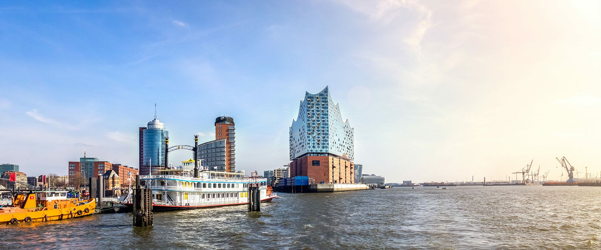 Panoramablick auf Hamburgs Hafen mit Elbphilharmonie, modernen Gebäuden, einem Raddampfer und weiteren Booten unter blauem Himmel.