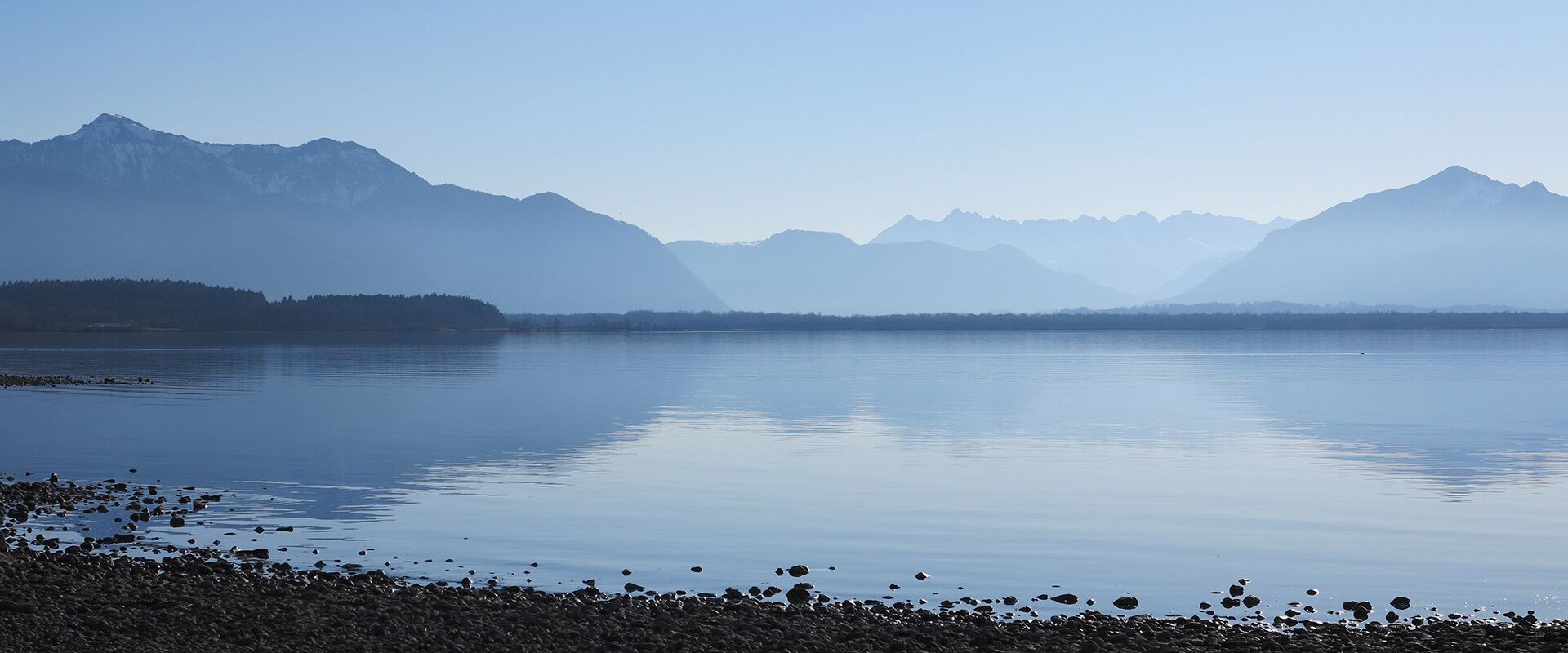 Panoramablick auf einen ruhigen See mit bewaldetem Ufer und gestuften blauen Bergen unter klarem Himmel.