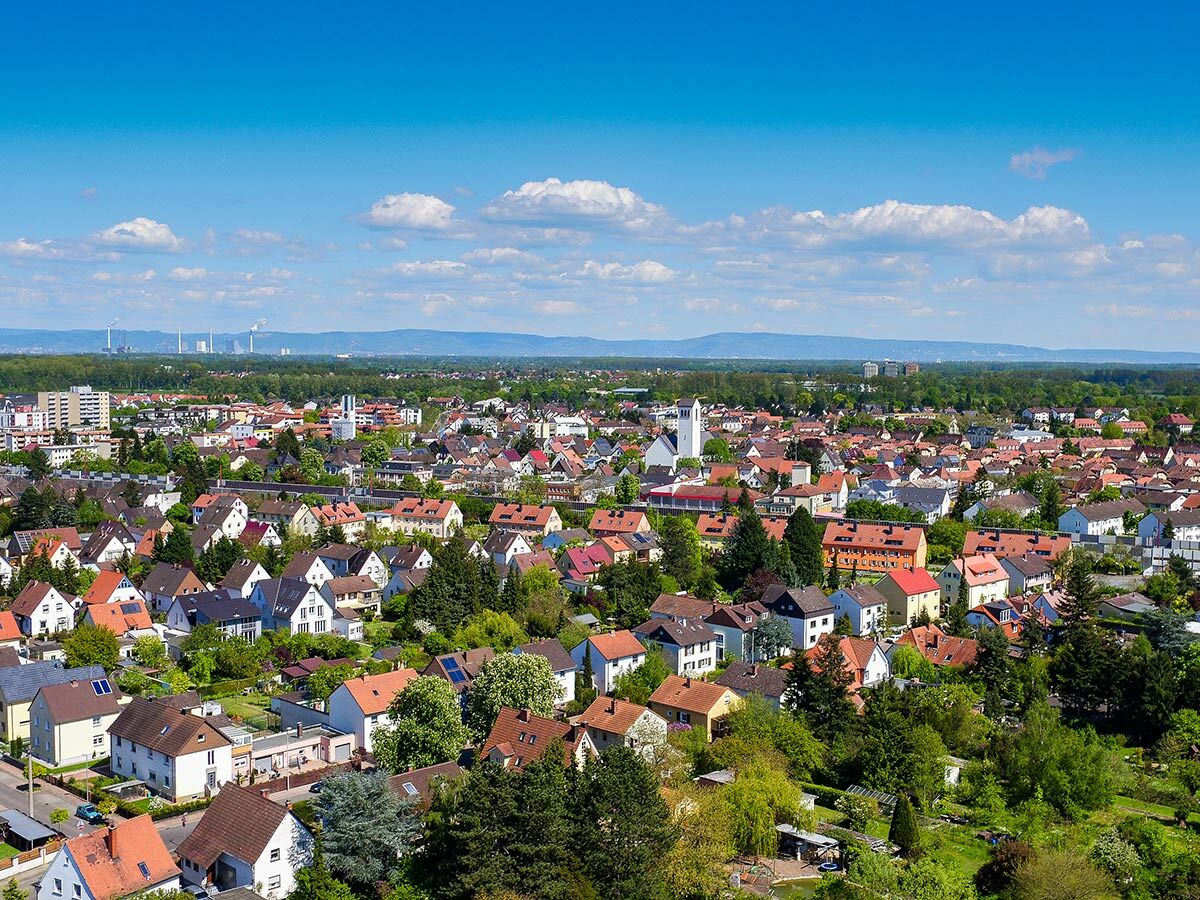 Luftbild einer deutschen Wohnsiedlung mit vielen Häusern, roten Dächern, grünen Gärten und Bäumen unter blauem Himmel. Industrieanlagen am Horizont.