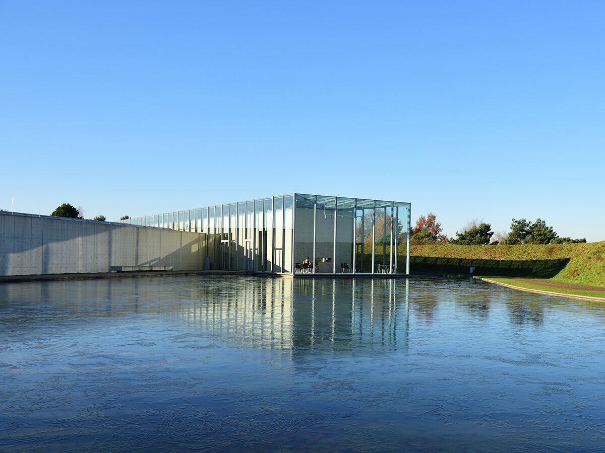 Modernes Gebäude mit Glasfassade und Betonwand, umgeben von Wasser und Grünflächen unter blauem Himmel. Architektur reflektiert.