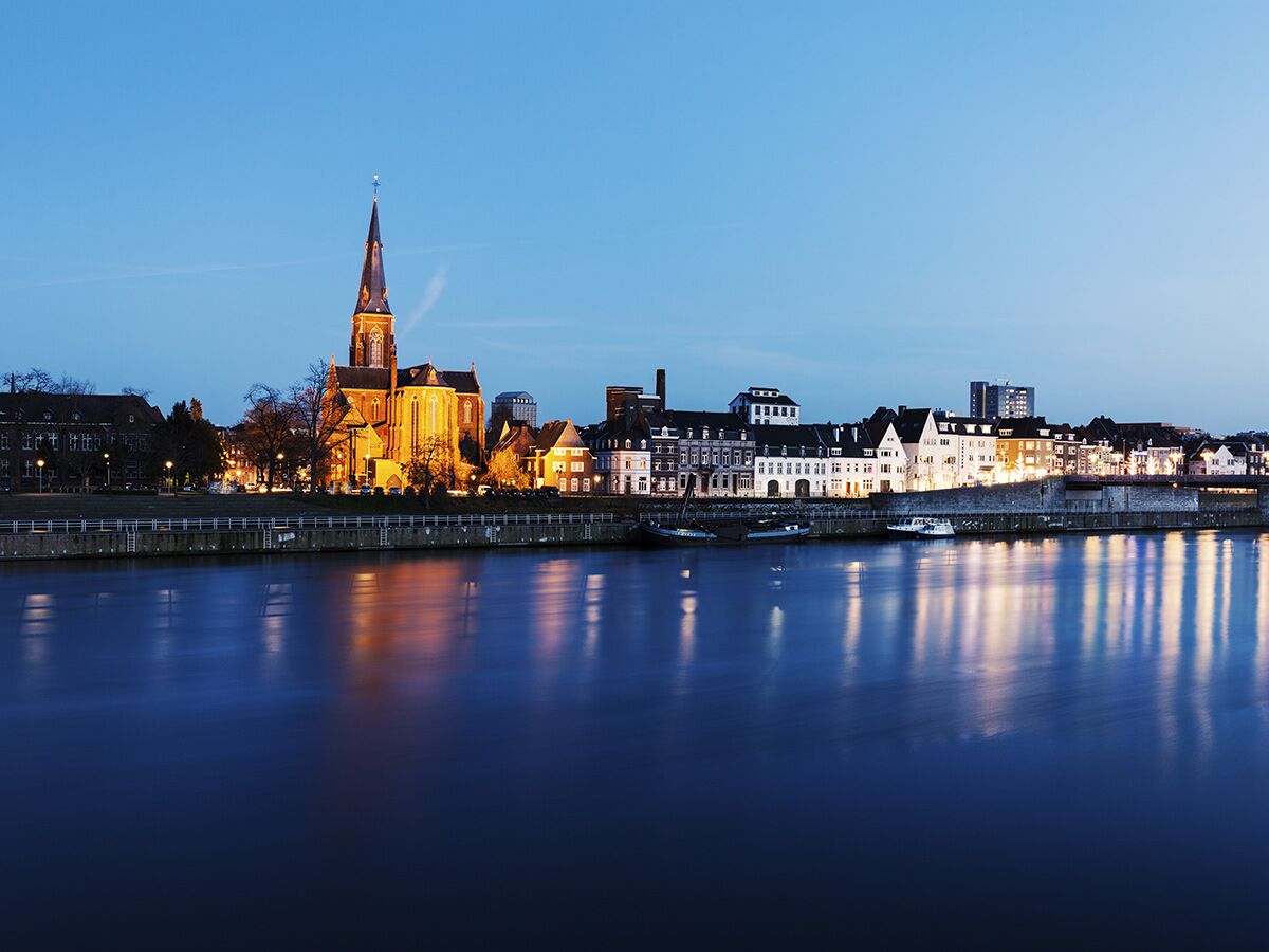 Abendliche Stadtansicht mit einem beleuchteten Flussufer, historischen Gebäuden und einer prominenten Kirche, die sich im Wasser spiegeln.