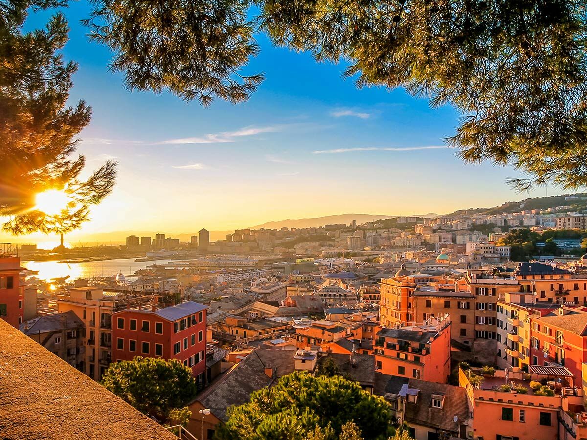 Panoramablick auf Genua bei Sonnenuntergang mit Hafen, historischen Gebäuden und Bergen im Hintergrund, gerahmt von Bäumen.