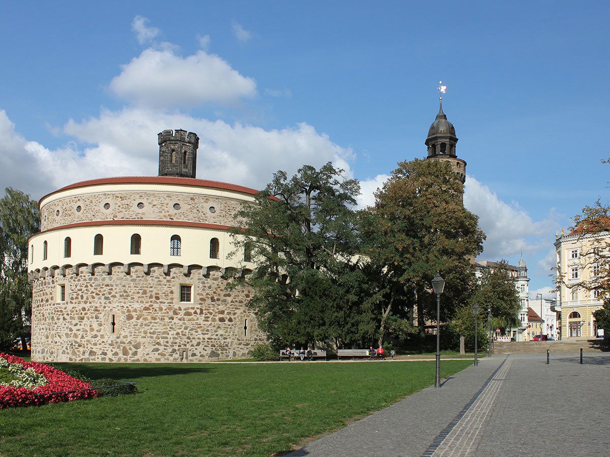 Ansicht des historischen Dicken Turms in Görlitz mit markantem Rundbau, Steinfassade und Zinnen auf sonnigem Stadtplatz.