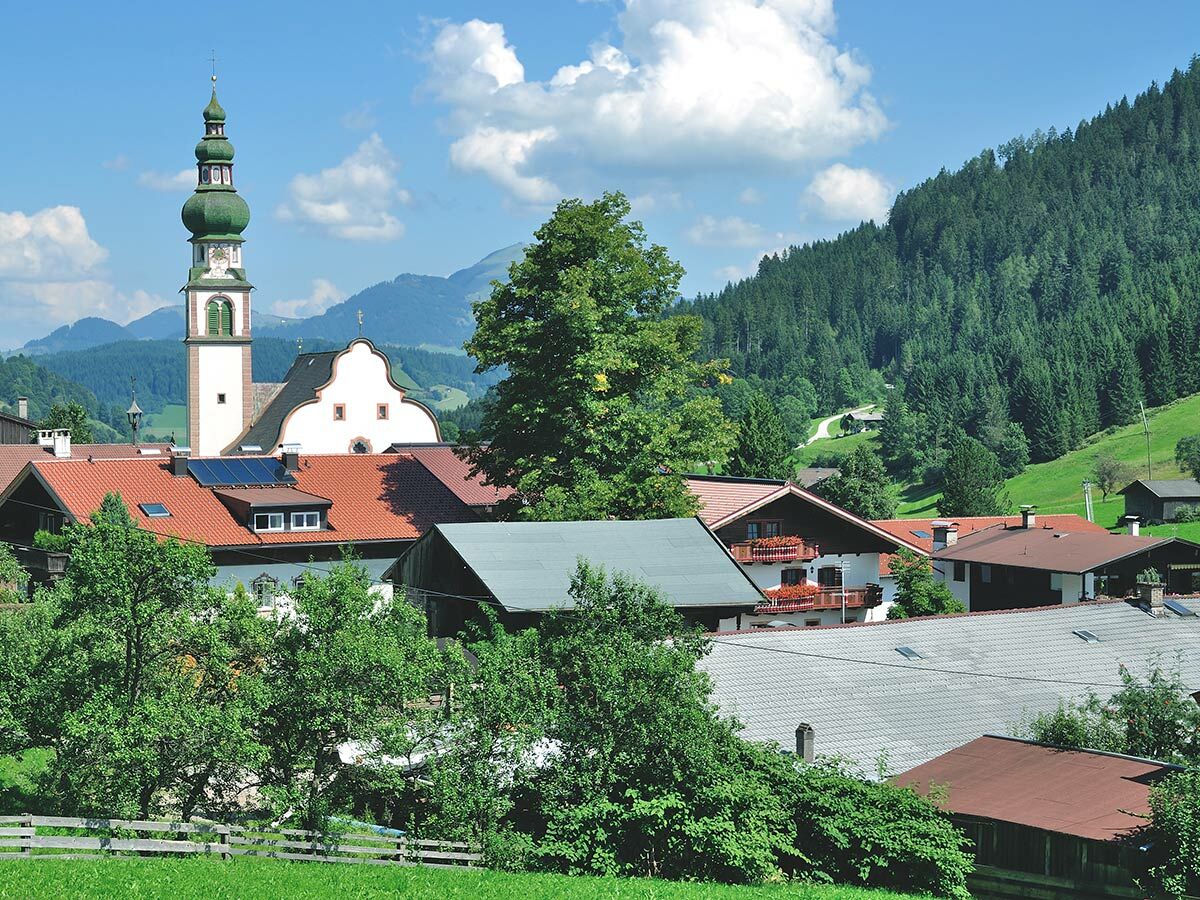 Blick auf ein malerisches Alpendorf mit traditionellen Häusern, Kirchturm mit Zwiebelkuppel, umgeben von grünen Wiesen und Wäldern.
