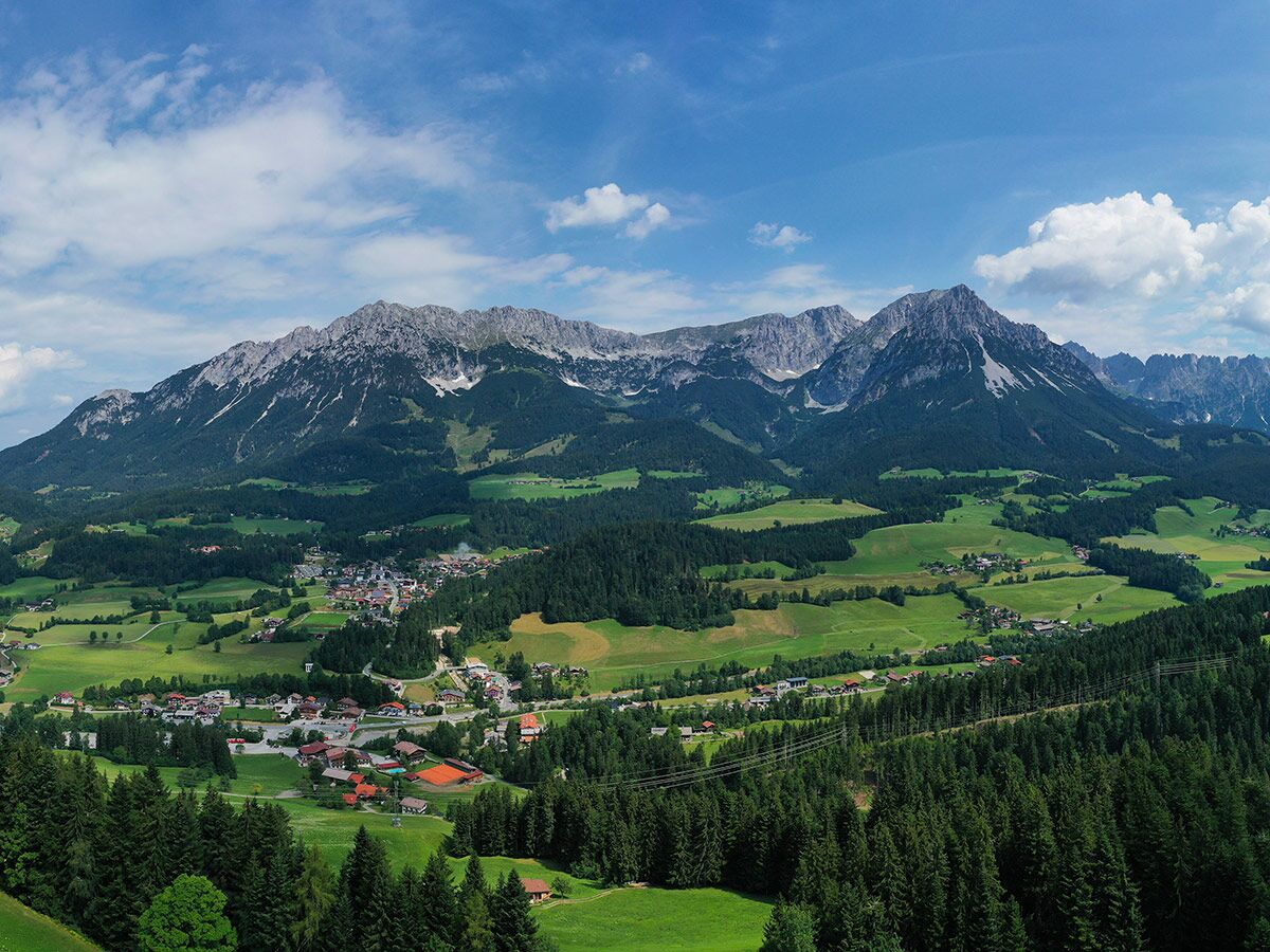 Panoramablick auf ein idyllisches Alpendorf in einem grünen Tal, umgeben von dichten Wäldern und majestätischen Bergen unter blauem Himmel.