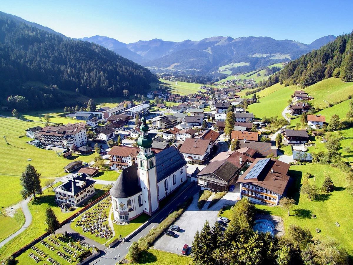Luftaufnahme eines idyllischen Alpendorfes mit markanter Kirche, umgeben von grünen Wiesen, Wäldern und Bergen unter blauem Himmel.