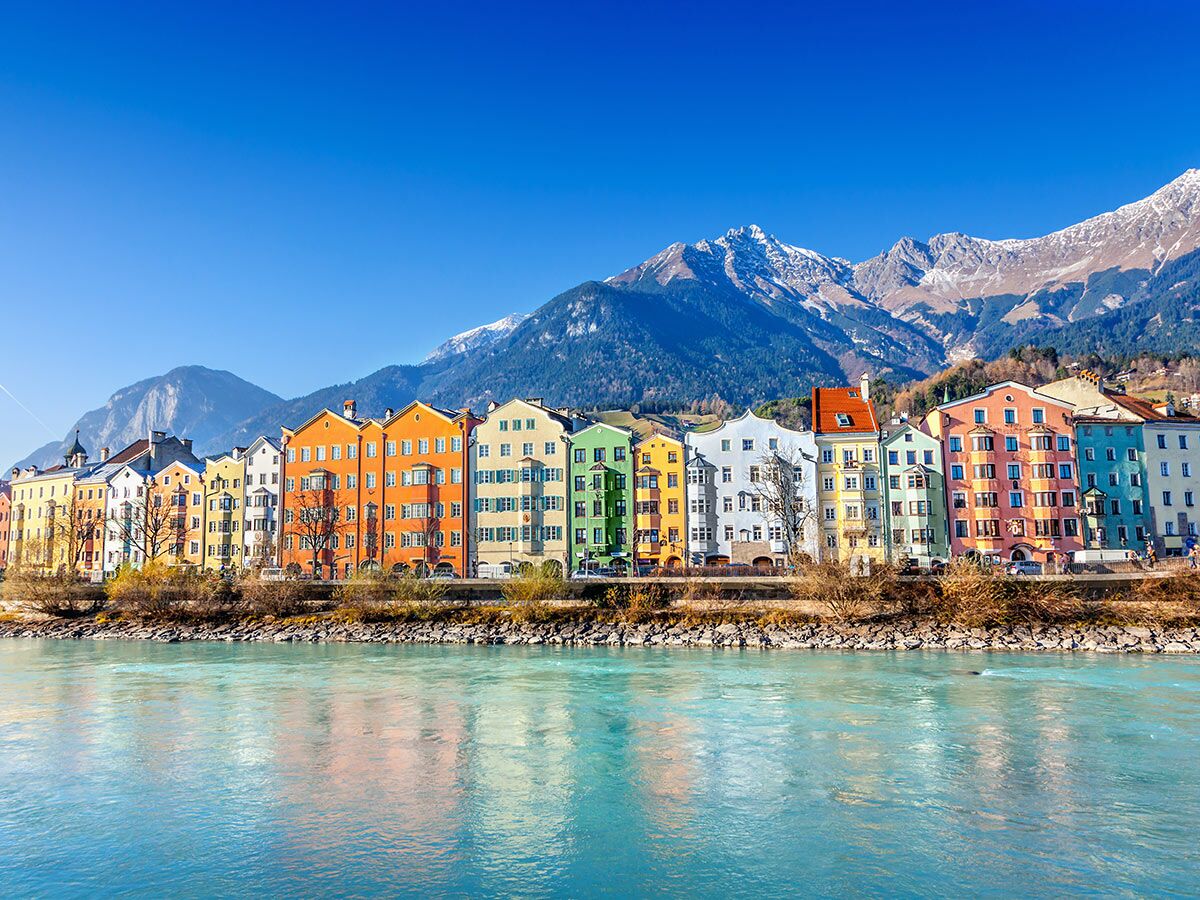 Panoramablick auf Innsbruck mit bunten Häusern entlang des Inn Flusses und schneebedeckten Alpen im Hintergrund.